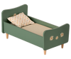 A small green wooden Maileg Wooden Bed, Mini with handpainted floral decorations and charming bunnies on the headboard and footboard. The bed has a beige mattress with thin stripes and a white pillow with brown polka dots. It stands gracefully on four short wooden legs.