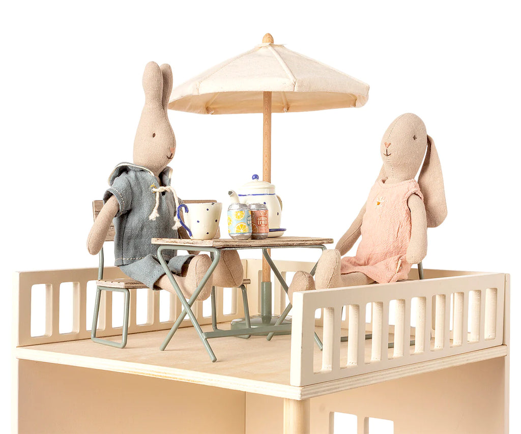 Two stuffed rabbits sit at a small table with a tea set, under a beige parasol. One rabbit wears a blue outfit, while the other wears pink. The Maileg | Miniature Garden Table Set, made from responsibly sourced wood, holds a teapot, cups, and jars on a small raised platform with a simple railing.