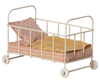 The Maileg Set - Sleepover At Grandparents features a miniature cream-colored metal baby crib with four wheels, ideal for imaginative play. It comes complete with a pink striped mattress, a yellow knitted blanket, and a pink pillow adorned with a floral pattern.