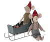 Two stuffed toy mice wearing red hats are positioned together. One mouse is seated on a Maileg Christmas Medium Sleigh - Green with a pair of striped mittens beside, while the other sits on the ground in front, wearing grey overalls and a plaid shirt adorned with hand-painted details.