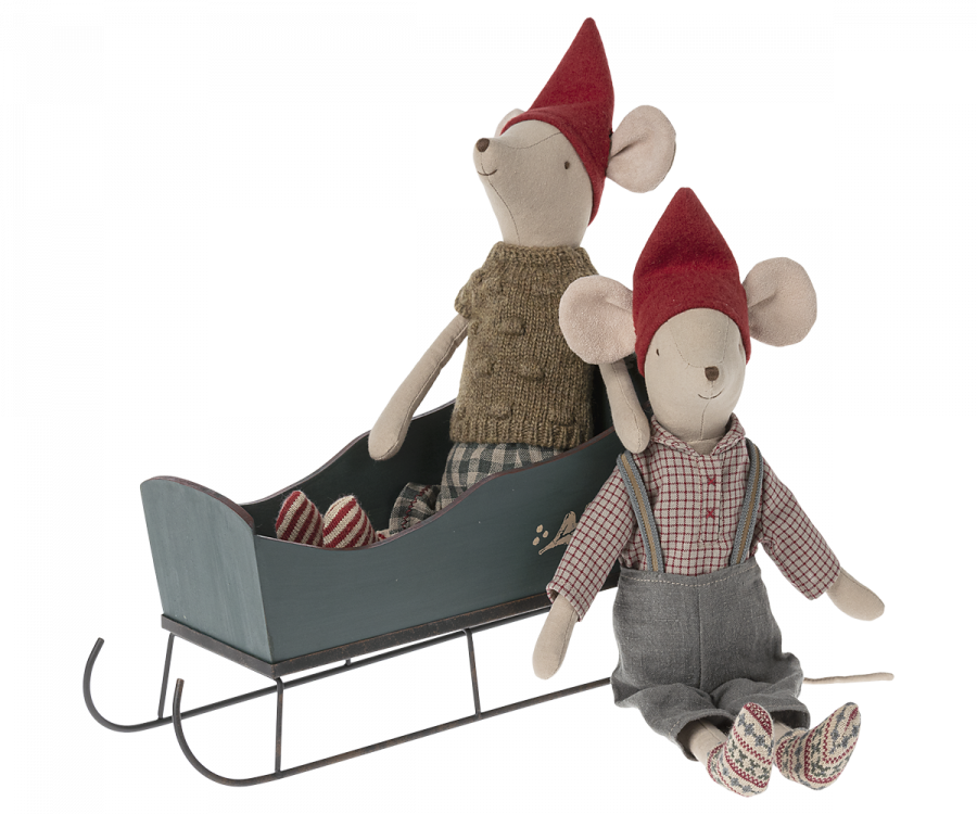 Two stuffed toy mice wearing red hats are positioned together. One mouse is seated on a Maileg Christmas Medium Sleigh - Green with a pair of striped mittens beside, while the other sits on the ground in front, wearing grey overalls and a plaid shirt adorned with hand-painted details.