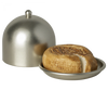 A Maileg Cheese Bell, a delicious surprise, is partially sliced and rests on a silver tray. A silver dome-shaped cover with a small knob on top is positioned to the left, slightly ajar to reveal the cheese inside. The cheese has a stamped design on the top, perfect for any mice treat.