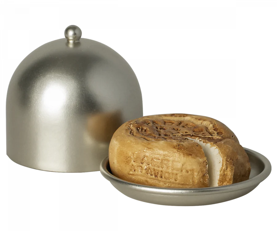 A Maileg Cheese Bell, a delicious surprise, is partially sliced and rests on a silver tray. A silver dome-shaped cover with a small knob on top is positioned to the left, slightly ajar to reveal the cheese inside. The cheese has a stamped design on the top, perfect for any mice treat.