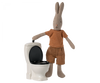 A plush rabbit wearing a striped shirt and brown shorts stands beside the Maileg Miniature Toilet - Teddy Size with the lid open. Part of the teddy family, the rabbit's arm rests on the toilet, as if gesturing toward it, creating a delightful scene in your bathroom assortment.