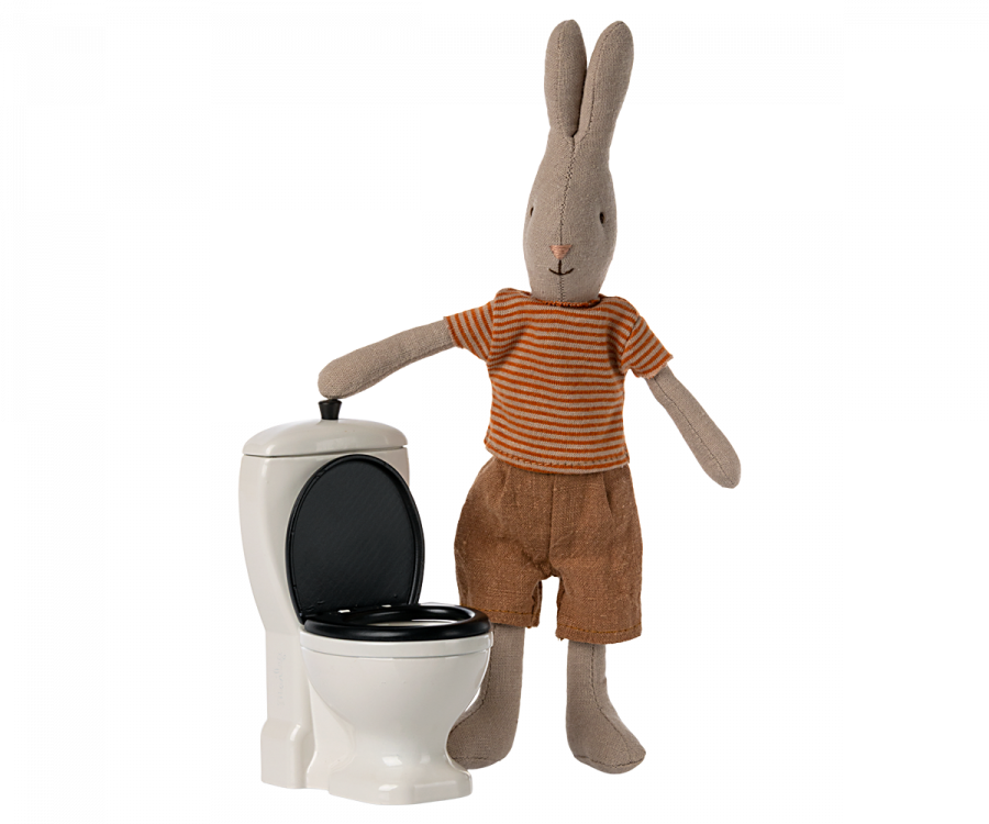 A plush rabbit wearing a striped shirt and brown shorts stands beside the Maileg Miniature Toilet - Teddy Size with the lid open. Part of the teddy family, the rabbit's arm rests on the toilet, as if gesturing toward it, creating a delightful scene in your bathroom assortment.