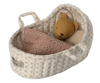 A small beige doll basket with tiny floral prints contains a brown stuffed bear tucked in with a soft, pink knitted blanket. This Maileg Carry Cot, Micro features fabric handles and a pocket on the side, making it perfect as a portable rabbit carrycot.