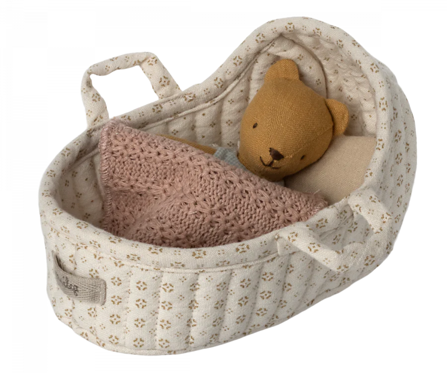 A small beige doll basket with tiny floral prints contains a brown stuffed bear tucked in with a soft, pink knitted blanket. This Maileg Carry Cot, Micro features fabric handles and a pocket on the side, making it perfect as a portable rabbit carrycot.