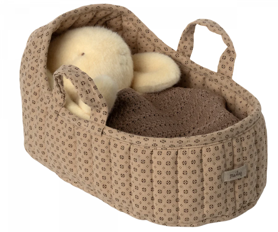 A plush teddy bear nestled inside a large, sand-colored Maileg Carry Cot, adorned with a knitted brown blanket. The carrycot features two convenient handles, making it perfect for cozy travels.