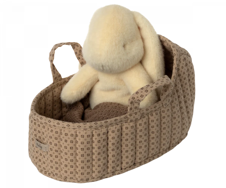 A plush bunny rests inside the Maileg Carry Cot, Large - Sand, featuring a charming quilted texture and convenient handles. The basket's patterned beige design resembles an adorable carrycot, complete with a small tag on the side. The bunny is crafted from a soft, off-white material.