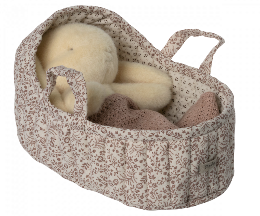 A plush teddy bear is cozy in a large, off-white Maileg Carry Cot with two handles, resembling a portable bed. A warm knitted blanket rests beside the bear.