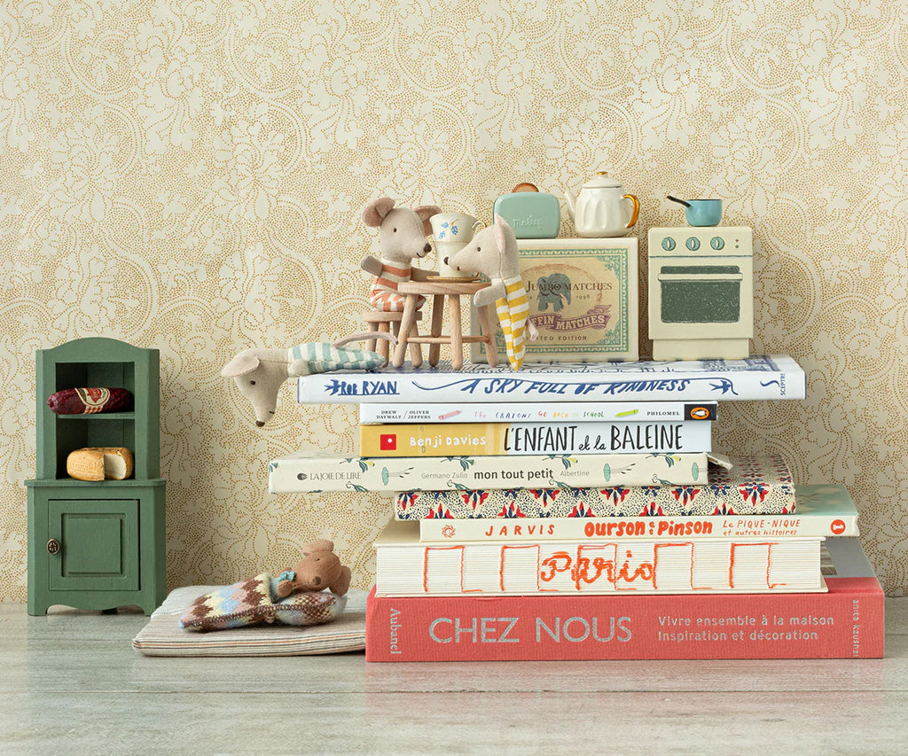 A whimsical scene features small mouse figurines seated at a table atop a stack of books. Nearby, the Maileg Corner Cabinet, Mouse - Dark Green adds charm with its vintage finish and practical storage space. The backdrop is a patterned beige wallpaper, complementing the cozy and imaginative home decoration.