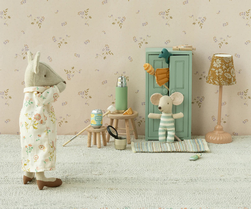 A small, plush rhinoceros in pajamas stands on a carpet, facing a plush mouse from the Maileg mice family dressed in striped clothes. The mouse is near the Maileg Mouse Closet in soft blue, adorned with golden hangers and topped with a toy carrot. The room features flowery wallpaper and a small floor lamp.