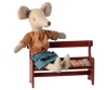 A charming plush mouse with a vintage flair is outfitted in an orange striped shirt and blue shorts. It relaxes on the Maileg Mouse Bench - Red, which includes a patterned seat cushion, with one arm draped casually over the back. Its large ears hint that it belongs to an adorable mouse family.