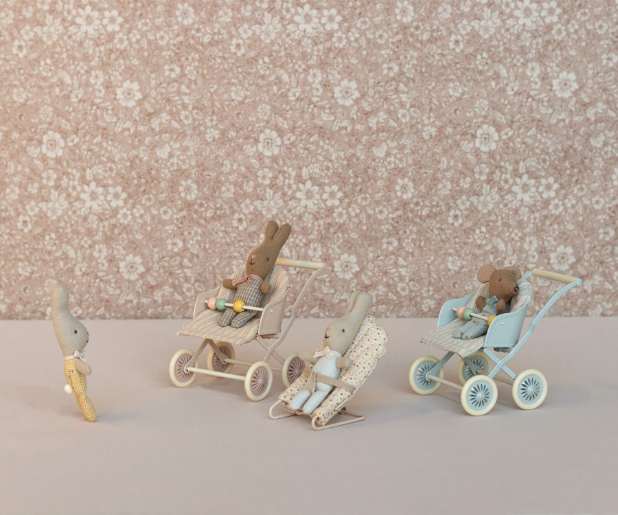 Three rabbit dolls, each in a Maileg Stroller, Baby Mice - Blue, are positioned on a floral patterned background. The strollers are light blue, beige, and blue with polka dot fabric. A fourth smaller rabbit doll walks towards them carrying a toy carrot, reminiscent of how mouse families transport babies.