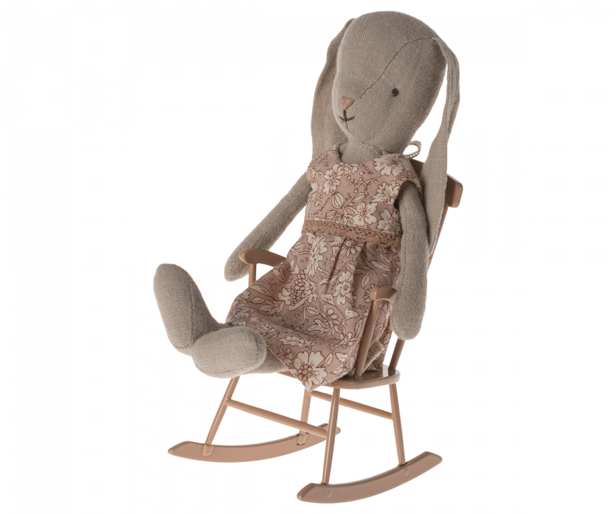 A stuffed rabbit toy wearing a floral-patterned dress is sitting in a small wooden rocking chair. Part of the teddy family, this bunny furniture piece features long ears and a neutral facial expression. The light brown Maileg Rocking Chair, Mini - Dark Powder (Larger Size) complements the brown and white floral design of the rabbit's dress.