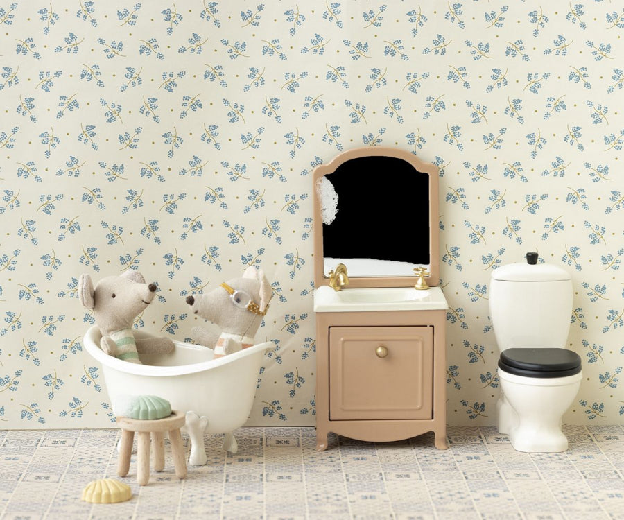 A miniature bathroom setup featuring two toy bears in a small white bathtub. The background has floral-patterned wallpaper, and there is a small vanity with the Maileg Sink Dresser With Mirror, Mouse – Powder, alongside a toilet with a black seat. A sponge and duck toys can be seen on the floor.