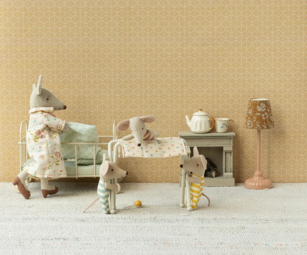 A miniature scene features a family of fabric mice in a cozy room. One adult mouse is by the crib, another rocks a baby mouse, and two child mice stand nearby in pajamas. The room includes miniature furniture, with the Maileg Nursery Table for Baby Mouse - Off White adorned with a tea set on it, and patterned wallpaper—a charming world for these baby mice.