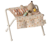 A small stuffed toy mouse lies on the Maileg Nursery Table, Baby Mouse - Off White, adorned in a striped pink and white onesie. Beside it, there is a green baby bottle and a yellow pacifier. The table cover features multi-colored polka dots, evoking the playful charm of a nursery table for baby mice.