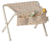 A small white baby bassinet with a polka dot patterned quilt. The quilt has small pockets on its side, one containing a baby bottle and the other containing a baby rattle. Nearby, the Maileg Nursery Table in off white for a baby mouse is thoughtfully arranged with essentials for easy access. The bassinet legs are crossed for support.