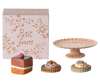 A decorative box labeled "Petit Four" is placed next to a pink, fluted pedestal cake stand adorned with teddy family accessories. In front are three small, intricately decorated pastries, each featuring unique toppings and designs, making up the charming Maileg Miniature Cakes & Cakestand set (ships in approximately one week).