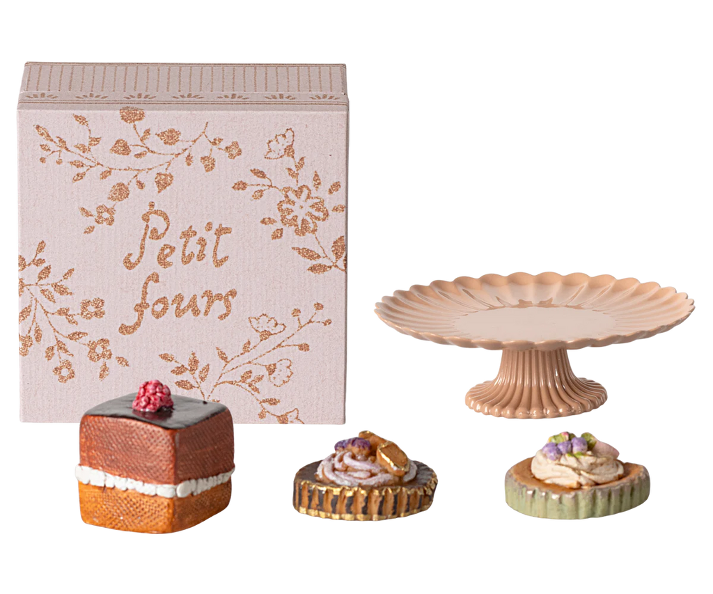 A decorative box labeled "Petit Four" is placed next to a pink, fluted pedestal cake stand adorned with teddy family accessories. In front are three small, intricately decorated pastries, each featuring unique toppings and designs, making up the charming Maileg Miniature Cakes & Cakestand set (ships in approximately one week).