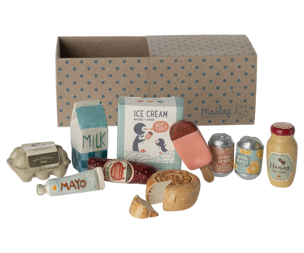 A delightful Maileg Miniature Grocery Box is displayed in front of a cardboard box with blue polka dots. Visible items include milk, ice cream, a popsicle, cans of sparkling water, a honey jar, a baguette, cheese, mayonnaise, an egg carton, and a jar of jam—perfect for any Maileg friend food collection.