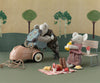 Two Maileg Picnic Set, Mouse Size plush toys in clothing are staged in a diorama. One mouse is examining a toy car's engine, while the other sits on a bench enjoying a drink. There's a picnic setup with food items on a checkered cloth nearby, along with a miniature fridge. The backdrop features stylized trees.