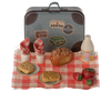 A small vintage-style suitcase, perfect as a dollhouse accessory, is adorned with travel stickers and sits on a checkered cloth. In front are two plates with bread, a loaf of seeded bread, two tomatoes, a small bottle of juice, and two napkins inside cups, arranged neatly for a picnic. The Maileg Picnic Set, Mouse Size completes this charming scene.