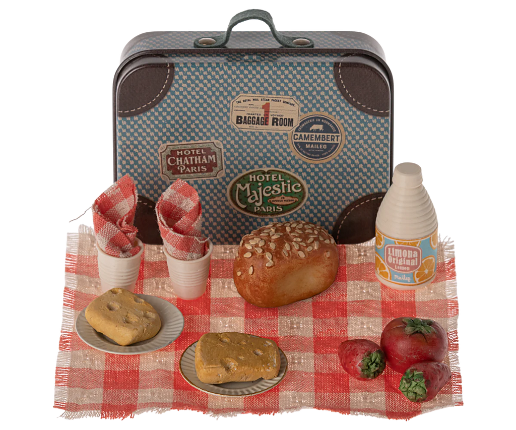 A small vintage-style suitcase, perfect as a dollhouse accessory, is adorned with travel stickers and sits on a checkered cloth. In front are two plates with bread, a loaf of seeded bread, two tomatoes, a small bottle of juice, and two napkins inside cups, arranged neatly for a picnic. The Maileg Picnic Set, Mouse Size completes this charming scene.