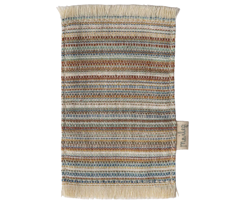 The Maileg Striped Rug, Small showcases a textured design with multicolored stripes and earthy shades of brown, green, and beige. It includes fringe on the top and bottom edges, making it perfect for use as a door mat in entryways. There is also a small tag attached to the right side.