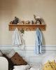 A cozy nook with a Ferm Living Large Place Rack - Oak holding various ornaments, a plush animal, and a wreath, above hooks with hanging towels and a robe. Decorative pillows are partially seen on a sofa below.