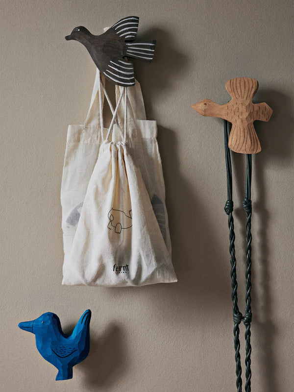 A canvas drawstring bag with a simple geometric design hanging on the Ferm Living hand-carved wooden bird hook, flanked by colorful bird-shaped wall decorations in black and blue, against a neutral-toned backdrop.