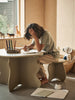 A child sits at a small Ferm Living Slope Table - Cashmere, concentrating on drawing in a well-lit kids' room with wood interiors. They hold their head thoughtfully while surrounded by colorful pencils and scattered paper.