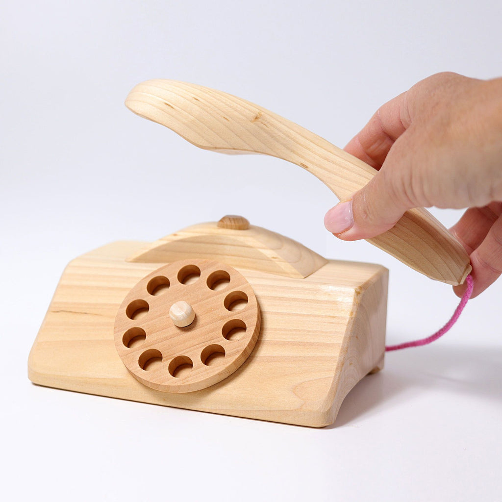 A person holds the receiver of the Grimm's Wooden Telephone, showcasing its light wood finish. This non-toxic toy features a circular rotary dial and a pink string connecting the receiver to the base, set against a plain white background.