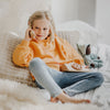 A young child with blonde hair sits on a cozy bed adorned with white and beige pillows, holding a Grimm's Wooden Telephone to their ear. Dressed in a yellow sweatshirt and blue jeans, the child appears engrossed in an imaginative conversation. Nearby, a stuffed animal and non-toxic toys are scattered playfully.