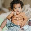 A toddler with curly hair, wearing an orange long-sleeve shirt and gray pants, sits on a bed holding Grimm's Wooden Telephone to their ear. Surrounded by soft, neutral-toned pillows and a non-toxic toy nearby, the child gazes upward with a curious expression.