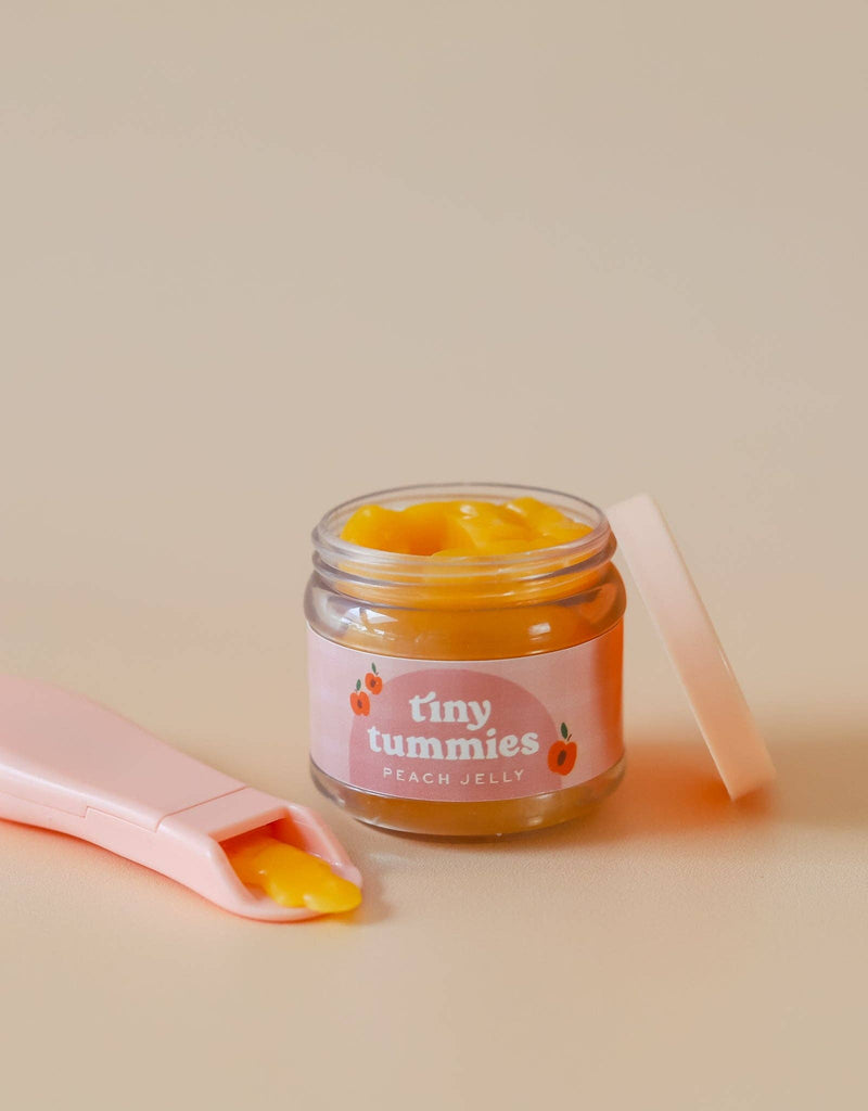 An open small glass jar labeled "Tiny Tummies - Peach Jelly Food" reveals a peach-colored jelly inside. Nearby, a pink magic spoon holds a small dollop of jelly. The background is soft and neutral in tone.