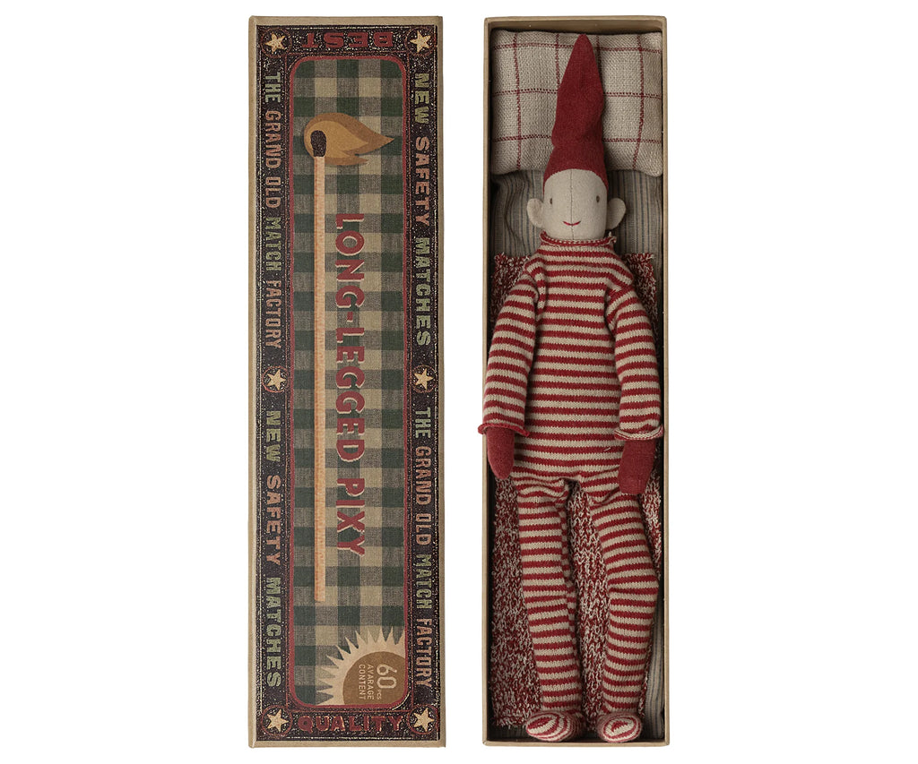 A red and white striped long-legged pixie doll wearing a knitted suit and a pointed red hat lies in a vintage-style decorative matchbox. The matchbox features elegant text and patterns, reading "The Grand Old Match Factory" and "Long-Legged Pixy," among other phrases, capturing the whimsical essence of Maileg Long Legged Pixy.