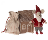 A small Maileg Santa Mouse, dressed in festive Santa clothes with a red hat, red and white outfit, and grey pants, stands next to a brown cardboard gingerbread house. The house features white icing-like decorations including a window and door. Nearby is a drawstring bag with red stitching—perfect for Christmas night.