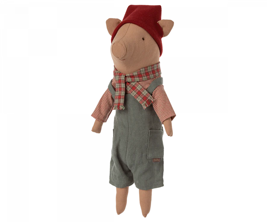 The Maileg Christmas Pig - Boy is a snuggly toy dressed in a red and white striped shirt, grey overalls with pockets, a red and green checkered scarf, and a matching red hat. Perfect for holiday decoration, the pig stands upright facing forward and features a removable hat for added fun.