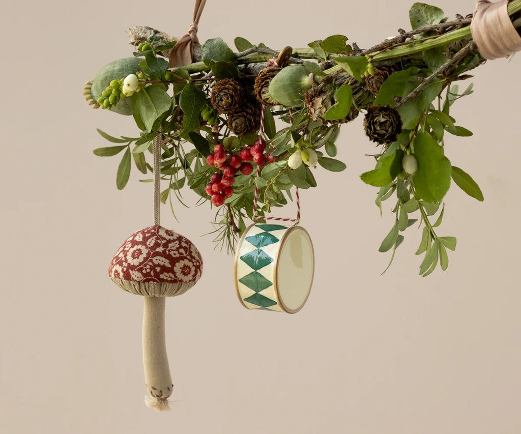 A decorative branch embellished with green leaves, pinecones, and red berries includes a charming fabric mushroom ornament with a patterned cap and a round ornament showcasing handpainted details in a geometric teal and white design. It's perfectly complemented by whimsical Maileg Metal Ornament, Large Drum - Dark Green.