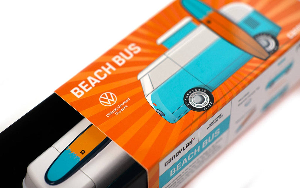 A toy box features a stylized illustration of a blue and white Volkswagen Type 2 van equipped with an orange surfboard on its roof. The box displays the name "VW Beach Bus Ocean" along with a Volkswagen licensed product logo, with the magnetic camper top toy partially visible inside the packaging.