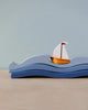 A small wooden toy sailboat with a white sail and orange hull is nestled between three stacked blue wave pieces, creating the illusion of the boat sailing on water. The background is a plain grayish-blue, and the scene is set on a light wooden surface, inviting open-ended play with imaginary sea creatures. Introducing the Blue Water and Sail Boat Set.