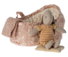 A small, plush Maileg Rabbit in Carry Cot, Micro - Rose wearing a knitted striped outfit sits on a cushion. Behind the rabbit is a pink floral-patterned basket with a knitted blanket draped over the edge. The charming playset is perfect for children and features a basket lined with light, patterned fabric.