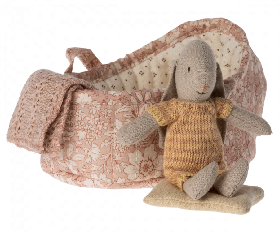 A small, plush Maileg Rabbit in Carry Cot, Micro - Rose wearing a knitted striped outfit sits on a cushion. Behind the rabbit is a pink floral-patterned basket with a knitted blanket draped over the edge. The charming playset is perfect for children and features a basket lined with light, patterned fabric.