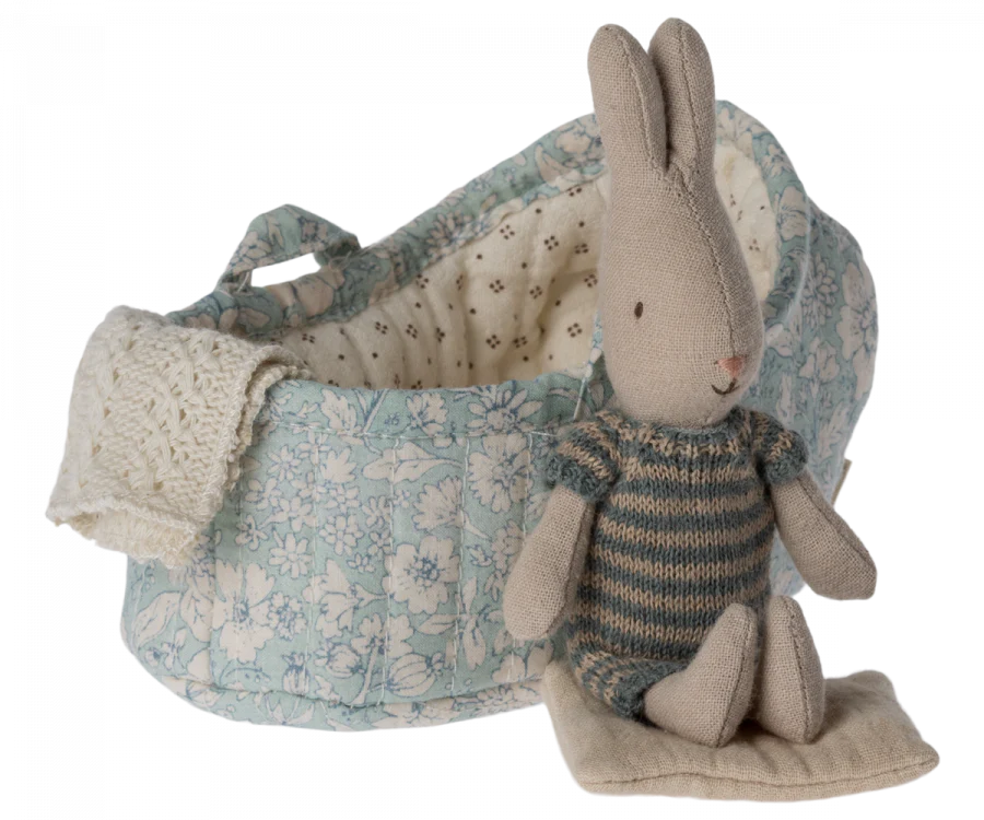 A Maileg Rabbit in Carry Cot, Micro - Blue sitting inside a floral-patterned miniature carry cot with a knitted blanket. The setting is isolated on a white background.