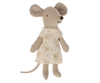 A Maileg little sister mouse wearing a beige Nightgown, standing upright against a black background. The mouse has large ears, a long tail, and a soft, textured body.