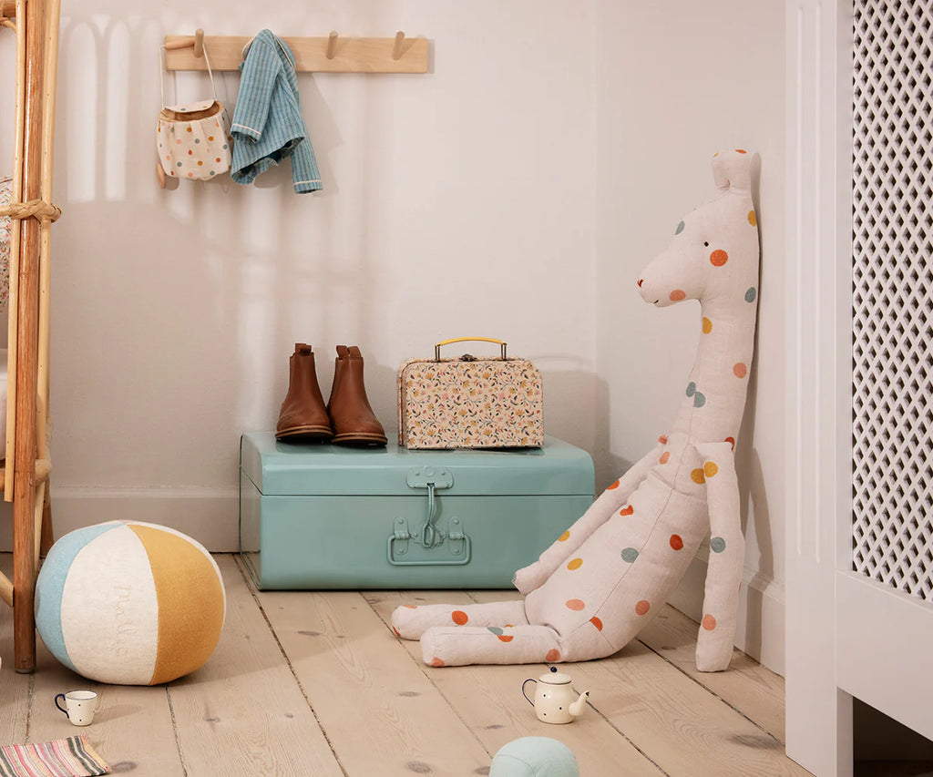 A cozy children's room showcases a pastel theme, highlighted by the Maileg Large Giraffe toy with its elongated body resting next to a teal chest. On the chest sit a floral suitcase and brown boots, while a colorful ball and toy cups are scattered on the floor. The wooden coat rack displays clothing and a beige bag, ready to welcome any safari friend.