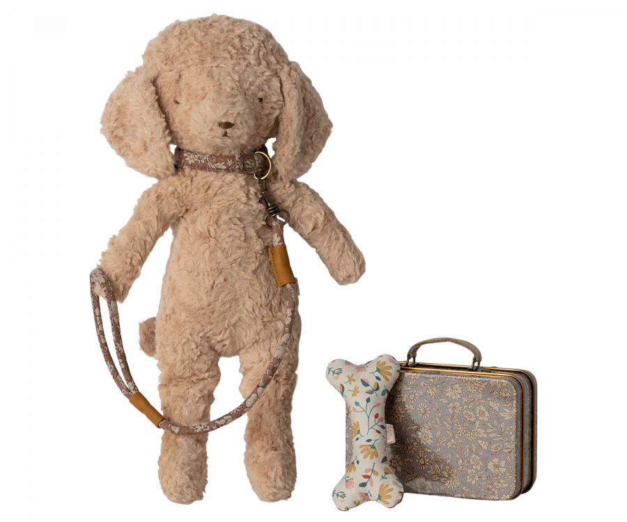 A Maileg Puppy Accessories - Rose with floppy ears stands upright, attached to a collar and leash. Beside the toy is a small metal suitcase and a white plush bone decorated with a floral pattern.