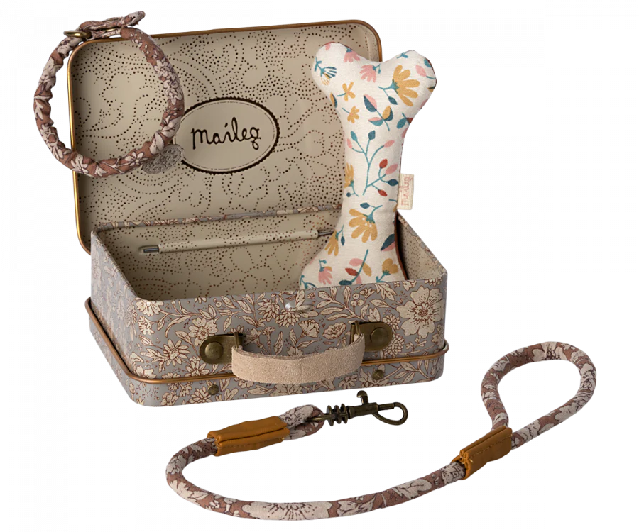 A small, metal suitcase with a floral pattern houses a plush dog toy shaped like a bone and adorned with vibrant flowers. Next to it are a matching floral-patterned collar and leash, perfect for pets. The suitcase cover features the brand name "Maileg Puppy Accessories - Rose" printed inside.